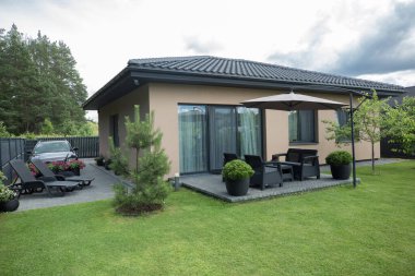 Bej duvarları ve koyu kiremitli çatısı olan modern bir özel evin genel planı. Evin önünde bir çimen, siyah mobilyalı asfaltlı bir teras ve büyük bir bej güneş şemsiyesi var..