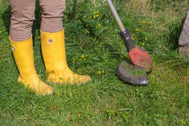 Sarı lastik çizmeli bir adam çimleri biçmek için elektrikli makina ya da fırça makası kullanır..