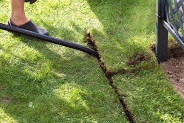 Fotoğraf küreğin üzerinde ayağı ile duran bir adamı gösteriyor. Küreği çimlere saplamaya çalışıyor. Sadece adamın bacakları, kürek ve çimlerin bir kısmı görülebiliyor..