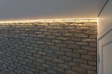 İç dekoratif tuğlayla kaplı bir aksan duvarlı oda. Duvarın üst kısmı LED şeritlerle aydınlatılmış, yumuşak ve sıcak bir ışık yaratıyor..