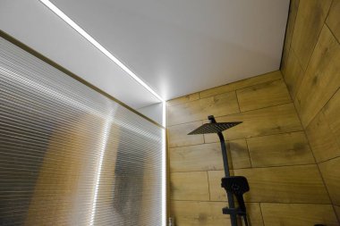 Sıcak tonların ve minimalist ışığın hakim olduğu bir duş. Duvarlar hafif ahşap desenli malzemelerle dolu ve tavan da LED şeritlerle donatılmış..