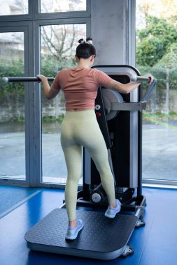 Genç sporcu kadın, modern bir spor salonunda gelişmiş spor aletleri kullanarak iç ve dış uyluk egzersizleri yapıyor. Geniş pencereler sayesinde geniş boşluklar doldurulabiliyor.