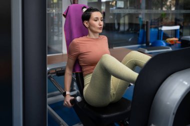 Spor salonunda ağırlık makinesiyle bacak çalıştıran genç sporcu kadın sırtında havluyu dinlendirirken ağırlık kaldırıyor, güç ve kas gelişimine odaklanıyor.