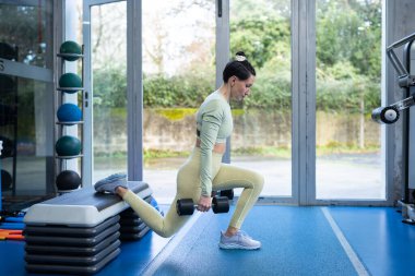 Genç atletik kadın denge ve destek için bir adım atarak spor salonunda dambıllarıyla ve kalça egzersizleri yapıyor.