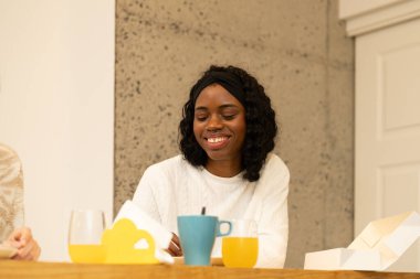 Kafasında saç bandı olan genç siyahi bir kadın modern mutfağında kahvaltı ederken gülümsüyor, portakal suyu, kahve, çay ve biraz hamur işi yiyor.