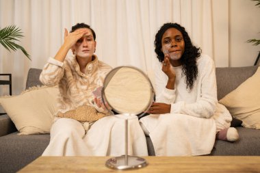 İki farklı bayan arkadaş evlerinde rahat bir akşamın tadını çıkarıyor, cilt kremlerinin bir parçası olarak nemlendirici yüz kremi sürüyorlar, rahatlama ve kendine bakmanın bir parçası olarak.