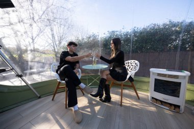 Romantik bir anın tadını çıkaran genç çift, doğal ortama dalmış özel bir glamping deneyimi için baloncuk çadırının içini görmek için kadehlerini kaldırıyor.