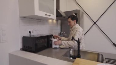 Genç bir kadın taze tost hazırlıyor, modern aletlerle kahvaltı hazırlarken rahatlamış sabah rutininin tadını çıkarıyor ve hoş bir gülümseme ile...