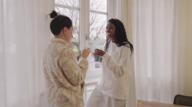 Rahat rahat giyinen iki farklı kadın, rahat bir evde pencerenin önünde durup, bardaklardan sıcak içecekler yudumluyor ve arkadaşça sohbet ediyorlar.