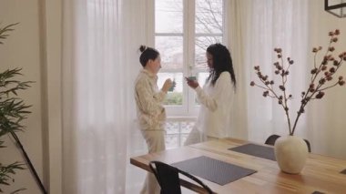 İki genç kadın, muhtemelen ev arkadaşları ya da arkadaşları, birlikte rahatlatıcı bir anın tadını çıkarıyorlar, çay yudumluyorlar ve rahat bir yuva ortamında parlak bir pencerenin önünde sohbet ediyorlar.