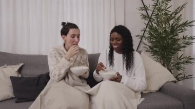 Battaniyeye sarılı genç kadınlar, oturma odasındaki kanepede film izlerken patlamış mısır paylaşıyorlar, yakın dostluk ve rahat bir aile anı sergiliyorlar.