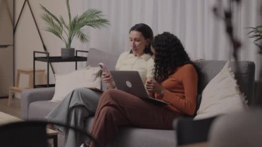 İki genç kadın, muhtemelen iş arkadaşları ya da ev arkadaşları, biri dizüstü bilgisayar, diğeri akıllı telefon kullanırken, modern bir oturma odasındaki koltukta rahatça oturuyorlar.