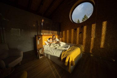 Genç bir çift, modern, çevre dostu bir otel odasında, rahat bir ortam yaratarak, dairesel bir pencereden içeri giren sıcak güneş ışığının tadını çıkarıyor.