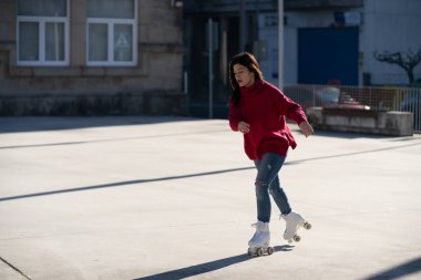 Young woman wearing a red sweater and ripped jeans roller skating in an urban environment, relishing the joy of rollerblading on a bright, sunny day clipart