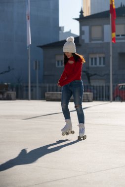 Kırmızı kazak giyen ve beyaz bereli genç bir kadın güneşli bir günde şehir ortamında paten kaymaktan hoşlanıyor. Gölgesi yerde süzülürken kayıyor.