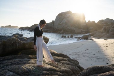 Gün batımında, şık plaj kıyafetleri içinde zarif genç bir kadın sakin ve tenha bir plajın kayalıklarında dikkatlice yürüyor, o anın huzurunun tadını çıkarıyor.