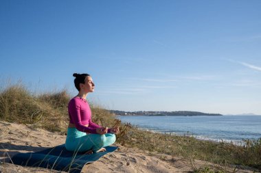 Yoga eğitmeni güneşli bir günde plaj minderinde meditasyon yapıyor. Arka planda deniz var. Farkındalığı ve sağlıklı yaşam tarzını teşvik ediyor.