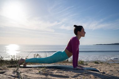 Spor giyim kuşamında genç bir kadın güneşli bir günde plajda yoga yapıyor, yukarı bakan köpek pozu veriyor, sağlık ve sağlığı destekliyor.