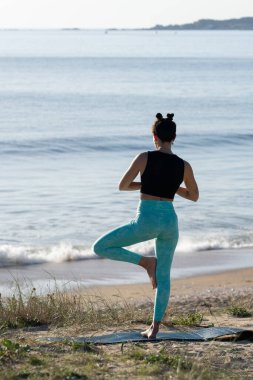 Spor kıyafetli genç bir kadın gün doğumunda plajda yoga yapıyor. Arka planda deniz varken ağaç pozu veriyor.