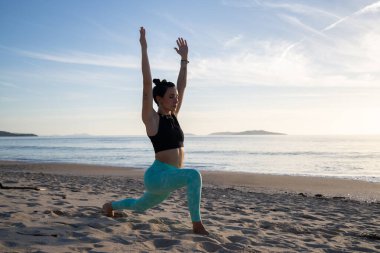 Dövmeli genç bir kadın kumlu bir plajda yoga pozu veriyor. Arka planda okyanus ve adalarla güneşin doğuşunun huzurunun tadını çıkarıyor.