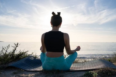 Kadın gün doğumunda plaj minderinde yoga ve meditasyon yapıyor. Okyanusun huzurlu atmosferini kucaklıyor ve o anda huzur buluyor.
