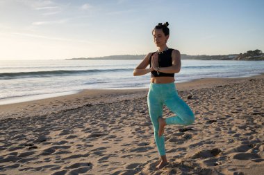 Concentrated yogini standing on one leg with hands joined in front of chest, performing vrikshasana on sandy beach with ocean waves at sunrise, promoting healthy lifestyle and mindfulness clipart