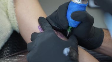 Profesyonel dövme sanatçısı, kadın müşterinin ön koluna dikkatlice kedi dövmesi çiziyor. Temiz ve iyi aydınlatılmış stüdyo ortamında çalışırken hassasiyet üzerine yoğunlaşıyor.
