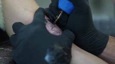 Profesyonel dövme sanatçısı koruyucu eldivenler takıyor. Dikkatli bir şekilde dövme makinesine rehberlik ediyor. Titizlikle müşterinin cildinde hassas ve dikkatli bir şekilde kalıcı bir tasarım yaratıyor.