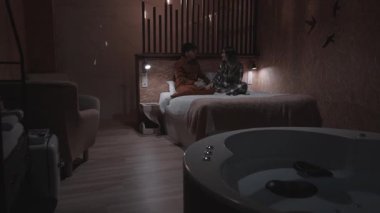 Otel odasındaki romantik akşam atmosferinde pijama giyen, el ele tutuşan ve yavaş çekimde sohbetin tadını çıkaran genç bir çift...