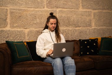 Evinden uzakta bilgisayarını kullanarak çalışan uzun saçlı genç adam modern bir dairede rahat bir kanepede oturmuş taş bir duvarla süslenmişti.