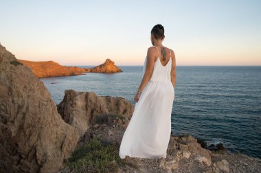 Beyaz elbiseli bir kadın Cabo de Gata doğal parkında bir uçurumun kenarında duruyor. Almeria, İspanya, Akdeniz ve volkanik kayaların üzerinde çarpıcı bir günbatımının tadını çıkarıyor.