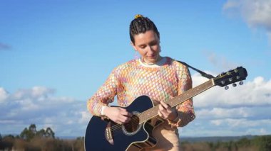 Profesyonel bayan müzisyen, kırsal manzaranın ortasında akustik gitar çalıyor. Geniş mavi gökyüzü altında canlı doğal ışık altında melodik sesler çıkarıyor.