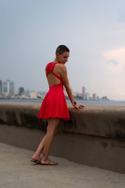 Canlı kırmızı elbise giyen genç bir kadın yıpranmış malecon duvarına yaslanmış dramatik havana şehri manzarasına ve bulutlu gökyüzünün altındaki geniş okyanusa bakıyor.