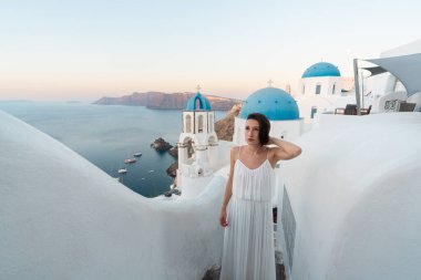 Oia, Santorini, Yunanistan 'da mavi kubbeli tipik bir beyaz yıkanmış kilisenin yanında duran genç bir kadın yaz tatilinin tadını çıkarıyor ve günbatımında Caldera manzarasına hayran.