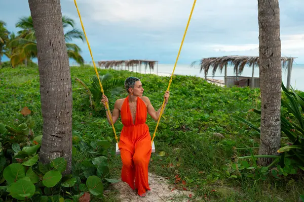 Kısa renkli saçlı, turuncu tulumlu, palmiye ağaçlarının arasında sallanan ve Cayo Guillermo, Küba 'da güzel playa pilar plajının keyfini çıkaran genç bir kadın.