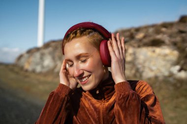 Gülümseyen sporcu kadın kablosuz kulaklıklarla müzik dinliyor. Arka planda rüzgar türbini var. Sürdürülebilir ve sağlıklı bir yaşam tarzını temsil ediyor.