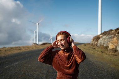Kahverengi eşofman giyen genç sporcu, mavi gökyüzü ve beyaz bulutlarla güneşli bir günde rüzgâr çiftliğinde kulaklıkla müzik dinlerken doğanın tadını çıkarıyor.