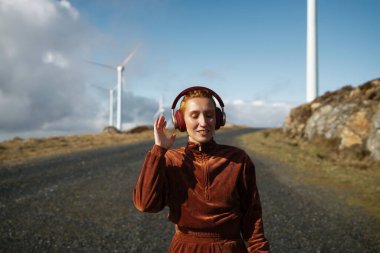 Genç, kızıl saçlı, kahverengi spor takım elbiseli, kulaklıkla müzik dinleyen, rüzgar çiftliği yakınlarında çakıllı bir yolda yürüyen, doğanın ve yenilenebilir enerjinin tadını çıkaran genç bir bayan.