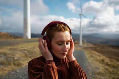 Kızıl saçlı sporcu kulaklığı kapalı kulaklıkla müzik dinliyor, rüzgar türbinleriyle manzaranın içinde duruyor, eğitimden sonra dinlenme anının tadını çıkarıyor.