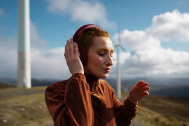 Kahverengi spor takım elbiseli kızıl saçlı sporcu rüzgâr tarlasında kulaklıkla müzik dinlerken güneşli bir günde temiz enerjiyi ve doğayla olan bağlantıyı hissediyor.