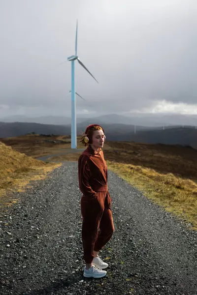 Kulaklık takan genç sporcu kadın, sürdürülebilir enerji ve yeşil yaşama adanmış pitoresk bir manzarada rüzgâr türbinleriyle çevrili çakıllı bir yolda dururken müziğin keyfini çıkarıyor.