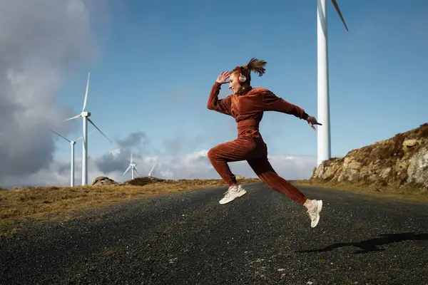 Kahverengi spor takım elbiseli ve kulaklıklı genç bir bayan, güneşli bir günde, rüzgâr türbinlerinin yakınındaki bir yolda zıplayarak müzik dinliyor.