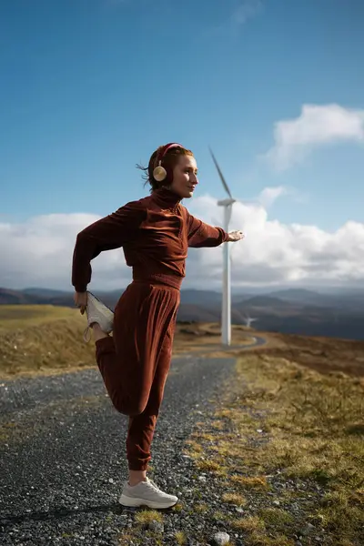 Kahverengi spor giyimli genç sporcu kadın güneşli bir günde bir dağ yolunda bacaklarını esnetiyor ve kulaklıkla müzik dinliyor. Doğanın tadını çıkarmak ve spor yapmak.