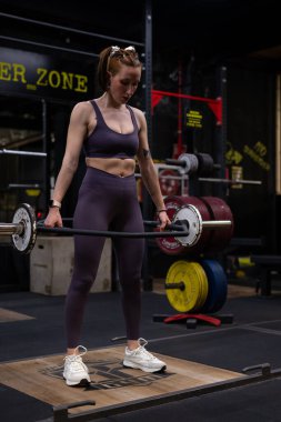 Genç bir kadın halterle kaldırma hareketini gerçekleştiriyor. Çağdaş bir jimnastik salonunda güç ve bağlılık gösteriyor. Aktif bir yaşam tarzının özünü somutlaştırıyor.