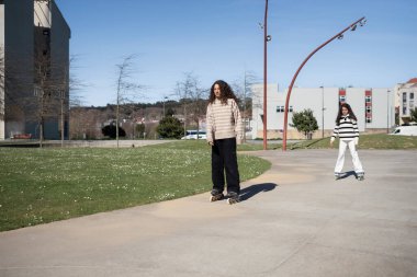 Güneşli bir şehir parkında babalarıyla paten kaymayı öğrenen iki genç kız, eğlenceli bir açık hava aktivitesi ve kaynaşma gününde neşe ve kahkaha paylaşıyorlar.