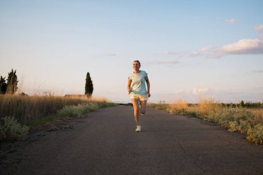 Kırsal kesimde kaldırımlı bir yolda koşan sağlıklı bir kadın, canlı günbatımının tadını çıkarırken dışarıda egzersiz yapmanın ve sağlıklı bir yaşam tarzını sürdürmenin keyfini çıkarıyor.