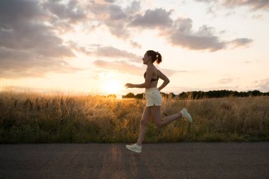 Genç bir kadın sağlıklı bir yaşam tarzını seviyor, gün batımında bir tarlanın yanındaki kaldırımlı yolda koşuyor, akşam antrenmanı sırasında doğanın sükunetini kucaklıyor.