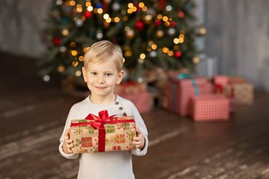 6 yaşında sarışın bir çocuk elinde bir Noel hediyesi tutuyor ve kameraya bakıyor. Arka planda hediyeli bir Noel ağacı var..