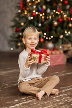 6 yaşında sarışın bir çocuk ahşap bir zeminde oturuyor, elinde bir Noel hediyesi tutuyor ve şaşırıyor. Arka planda hediyeli bir Noel ağacı var..