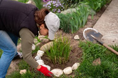 Kadın bahçedeki bir çiçek tarlasına dekoratif taşlar seriyor, köy hayatı kavramı.